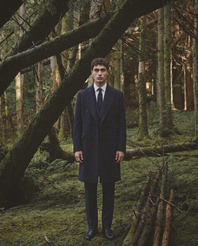 A man in a navy double breasted overcoat stands in a mossy forest.